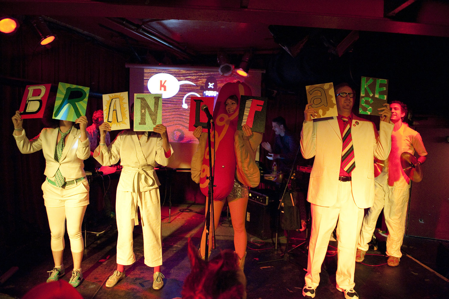 A photo from one of the Bran Flakes' live shows. There is a small group of people holding out signs that spell that name 'Bran Flakes'. There's also a lady in a hot dog suit.